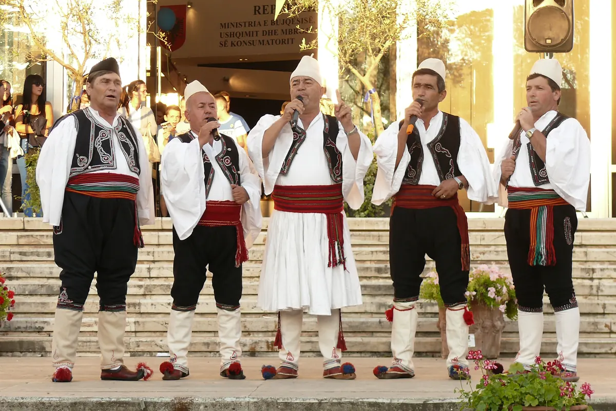 Five men in white shirts, embroidered waistcoats and striped sashes sing into microphones on an outdoor stage