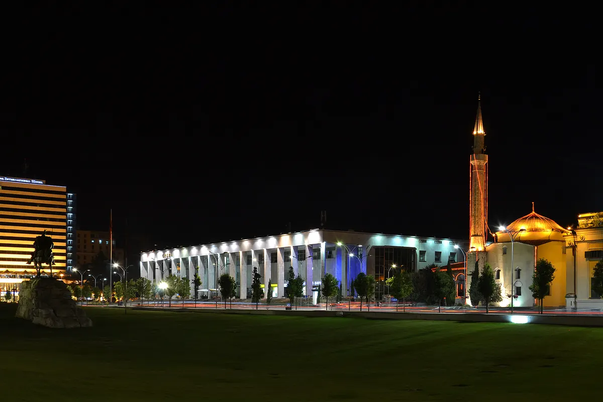 Skanderbeg Square at night, with a lit white colonnaded building at the centre, a lit hotel tower on the left and the golden dome and minaret of the Et'hem Bey Mosque on the right