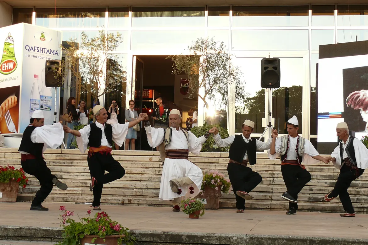 Men in white skirts, black waistcoats and felt caps dance hand in hand in a line on a stone stage with spectators behind them